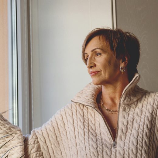 A trans woman dressed in a cream cable knit sweater is standing and looking out a window.
