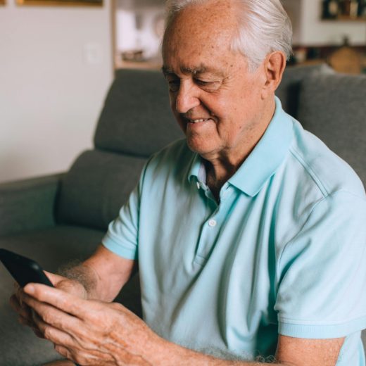 An elderly man dressed in shorts and a light blue polo t-shirt smiles as he uses his phone. He is seated ona green sofa.