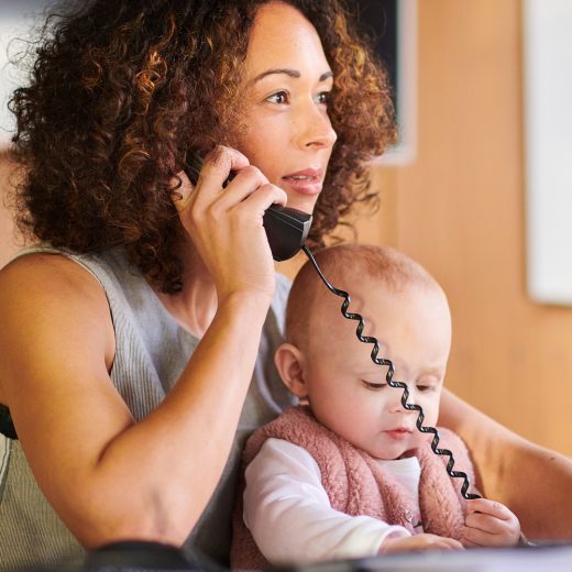 A woman with curly hair is using a landline telphone while a young baby sits on her lap.