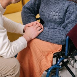 A man is down on one knee and is talking to a person in the wheelchair while he holds their hand.