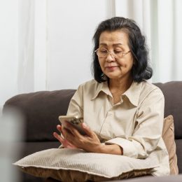 An older woman sits on a dark grey or brown sofa while looking at her phone. She is asian, has shoulder length dark hair and is wearing a cream shirt.