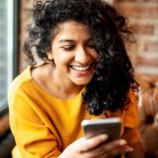 A young asian girl smiles happily as she looks at her phone, she has curly black hair and is wearing a yellow jumper.